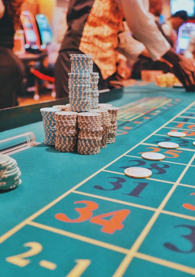 A casino scene with a roulette table features prominently, covered in green felt with colorful numbers and betting squares. Several stacks of poker chips are placed on different numbers, and the hands of several people are visible as they interact with the table. In the background, there are slot machines and more people, creating a vibrant and busy atmosphere.
