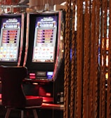 A pair of casino slot machines, each with detailed screens displaying various game outcomes and jackpot amounts. The chairs in front of the slot machines are dark and cushioned for comfort. The background features a hanging curtain made of golden ropes and other casino decorations are visible.