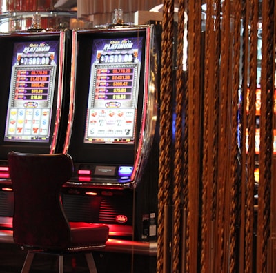 A pair of casino slot machines, each with detailed screens displaying various game outcomes and jackpot amounts. The chairs in front of the slot machines are dark and cushioned for comfort. The background features a hanging curtain made of golden ropes and other casino decorations are visible.