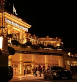 A luxurious casino at night, illuminated with warm, golden lights. The building features elegant architecture with columns and decorative elements. A few people are walking towards the entrance, and there are a couple of parked cars nearby.