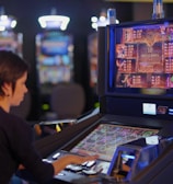 A person is seated at a slot machine console in a casino, focusing intently on the game displayed on the screen. The background shows several other illuminated gaming machines, adding a vibrant and colorful atmosphere to the scene.