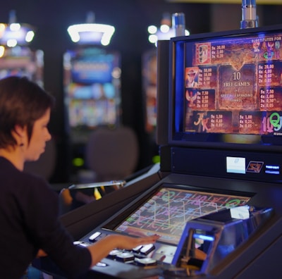 A person is seated at a slot machine console in a casino, focusing intently on the game displayed on the screen. The background shows several other illuminated gaming machines, adding a vibrant and colorful atmosphere to the scene.