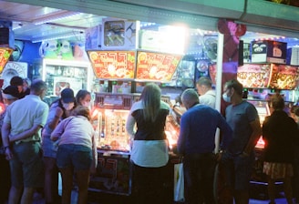 A group of people standing in front of an illuminated amusement arcade, engaging with various gaming machines. The environment is bustling with activity, and colorful lights create a vibrant atmosphere. The ceiling is decorated with large, playful images of dice and other game-related graphics, enhancing the lively mood.