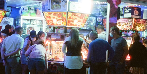 A group of people standing in front of an illuminated amusement arcade, engaging with various gaming machines. The environment is bustling with activity, and colorful lights create a vibrant atmosphere. The ceiling is decorated with large, playful images of dice and other game-related graphics, enhancing the lively mood.
