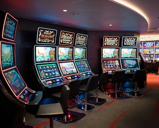 A row of brightly lit slot machines in a casino setting, each with colorful displays and various game themes. The machines are lined up against a dark wall and are unoccupied, with cushioned chairs placed in front of them. The area is dimly lit except for the glow from the screens, creating a vibrant and engaging atmosphere.