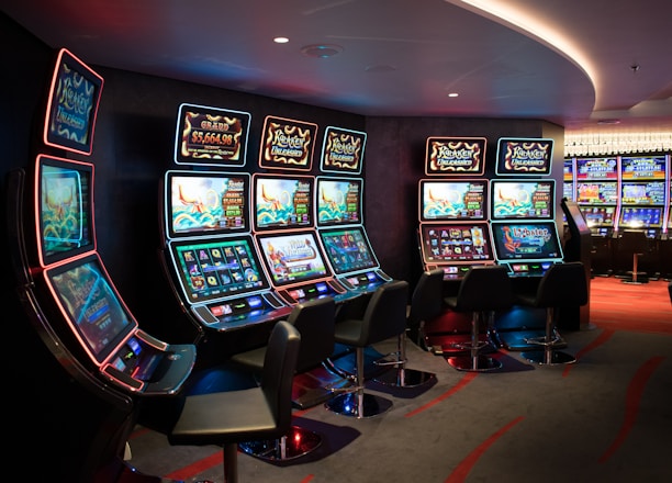 A row of brightly lit slot machines in a casino setting, each with colorful displays and various game themes. The machines are lined up against a dark wall and are unoccupied, with cushioned chairs placed in front of them. The area is dimly lit except for the glow from the screens, creating a vibrant and engaging atmosphere.