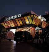 A building with a brightly illuminated entrance is seen at night. The sign above reads 'Casino' in bright letters. The facade features colorful graphics depicting various indulgent themes. Several vehicles are parked near the entrance, and people are visible in the vicinity. Tall lamp posts with spherical lights are present, casting a warm glow.