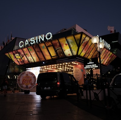 A building with a brightly illuminated entrance is seen at night. The sign above reads 'Casino' in bright letters. The facade features colorful graphics depicting various indulgent themes. Several vehicles are parked near the entrance, and people are visible in the vicinity. Tall lamp posts with spherical lights are present, casting a warm glow.