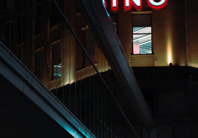 A night scene featuring a building with illuminated neon signage displaying 'CASINO'. The environment is urban with reflections on glass windows and glowing street traffic lights below. The atmosphere is dimly lit, with artificial lighting casting vibrant colors.